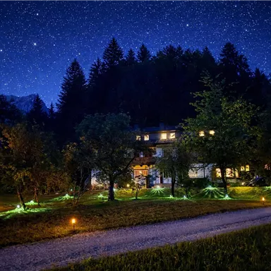 A picturesque building under a clear night sky filled with stars. The garden is illuminated and surrounded by trees.