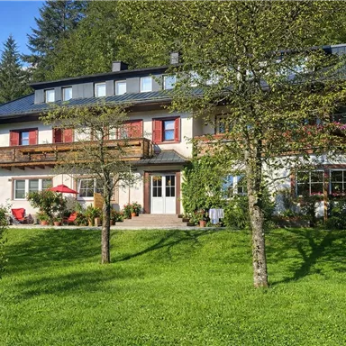 A charming multi-story house, surrounded by green grass and trees. In the background, there are tall mountains.