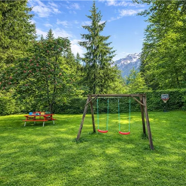 A beautiful garden with a swing and a picnic table. Surrounded by trees and a mountainous landscape in the background.