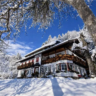 A picturesque winter house surrounded by snow-covered trees. The sky is clear and blue, creating a tranquil atmosphere.