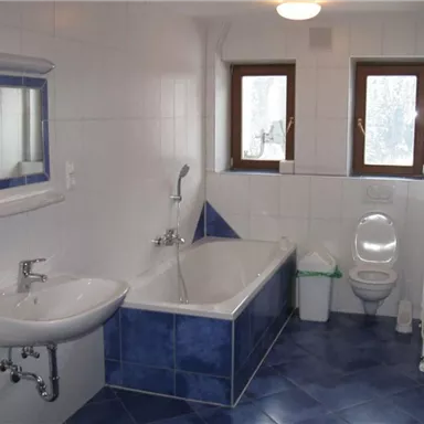 A bright bathroom with blue tiles and a bathtub. There is a sink, a toilet, and two windows for natural light.