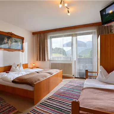 A cozy room with a double bed and a single bed.
Large windows offer a view of the mountains and there are wooden furniture as well as a television.