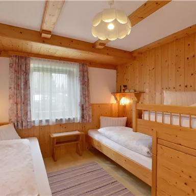 A bright room with wooden paneling and two beds, including a bunk bed. The room has a window with curtains and a cozy atmosphere.