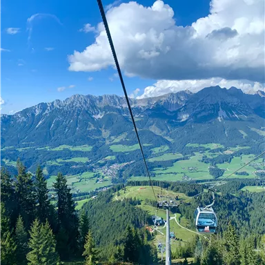 Eine Seilbahn fährt durch eine grüne Berglandschaft. Im Hintergrund sind majestätische Berge und ein blauer Himmel mit Wolken zu sehen.