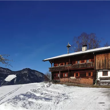 A cozy wooden house in the snow, surrounded by mountains. The clear blue sky creates a beautiful winter atmosphere.