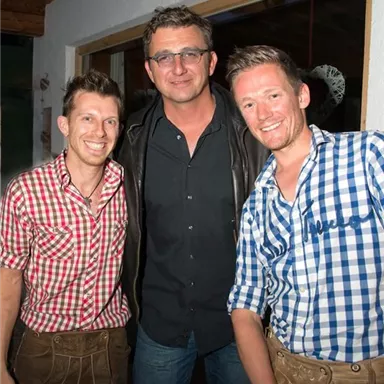 Three men pose together in front of a wall. They are wearing traditional costumes in checked patterns.