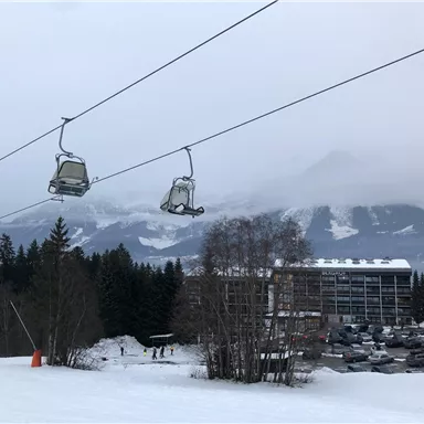 A ski lift system with chairs in a snow-covered landscape. In the background, there are mountains and a cloud cover.