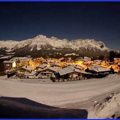 A peaceful mountain landscape at night with snow-covered rooftops and illuminated buildings. In the background, majestic mountains rise under a clear sky.