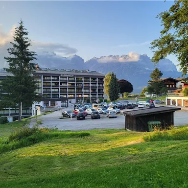 Ein großes Gebäude in den Alpen mit Parkplätzen im Vordergrund. Die Umgebung ist grün und von Bergen umgeben.