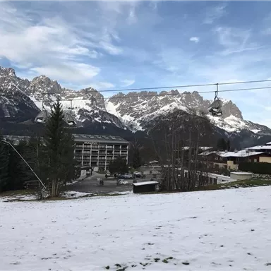 A snow-covered valley with majestic mountains in the background. In the foreground, you can see a hotel and a cable car.