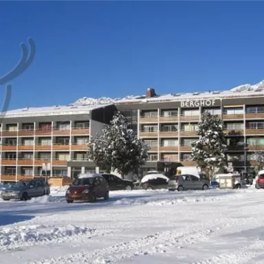 Ein Hotelgebäude im Schnee mit dem Schriftzug "Berghof". Umgeben von verschneiten Bäumen und einem klaren blauen Himmel.
