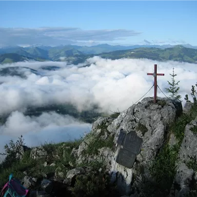 Ein Blick auf eine atemberaubende Berglandschaft mit Nebel und Wolken. Im Vordergrund steht ein Kreuz auf einem Felsen.