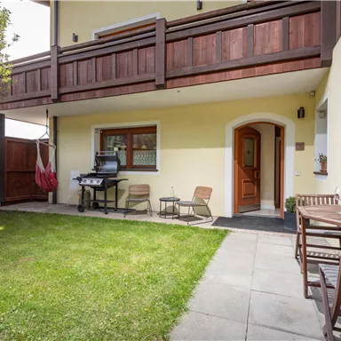 A cozy outdoor area with a small lawn, a grill, and wooden chairs. The surroundings are enclosed by a wooden fence and there is a hanging chair.