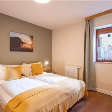 A cozy bedroom with a double bed and yellow pillows. The room has warm colors and a window with curtains.