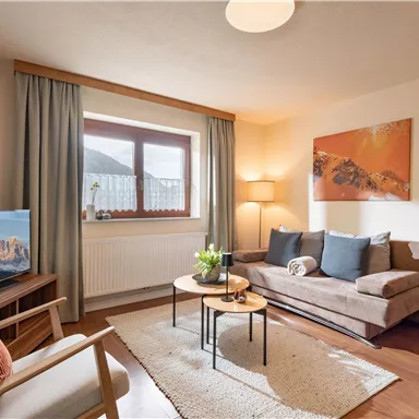 A cozy living room with a sofa, a coffee table, and a television. It has large windows and bright, friendly colors.