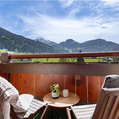 A cozy balcony with a wooden chair and table. In the background, green hills and mountains can be seen under a blue sky.