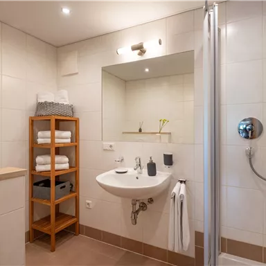 A modern bathroom with a shower, a sink, and folded towels. A wooden shelf with additional towels complements the space.