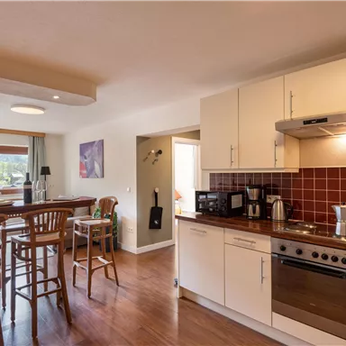 A modern kitchen with wooden flooring and bright furniture. In the background, there is a table with chairs and large windows that let in plenty of daylight.