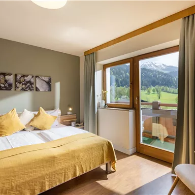 A cozy bedroom with a large bed and yellow pillows. The room has a window overlooking a green landscape and mountains.