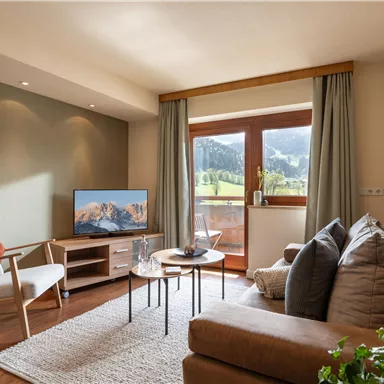 A cozy living room with a sofa, a TV, and a table. Large windows offer a view of the green mountains.