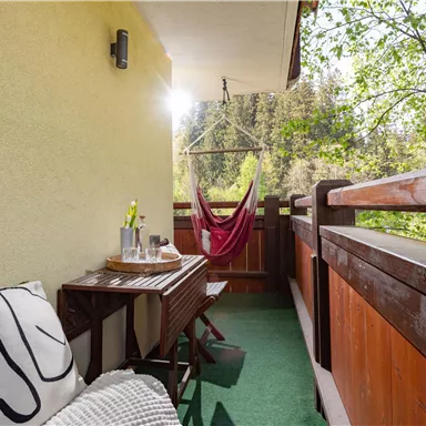A cozy balcony with a wooden table and a hammock. In the background, trees can be seen and the sunlight shines on the surface.