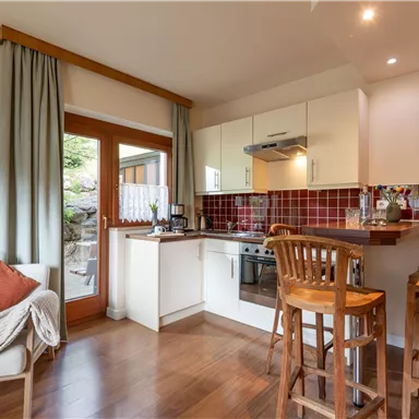 A modern kitchen with wooden flooring and red tiles. There is a breakfast bar with high stools and a view of the garden through the door.