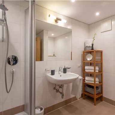 A modern bathroom with a shower, sink, and mirror. There is a wooden shelf with towels and decor.
