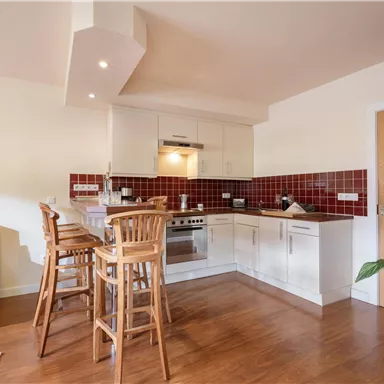 A modern kitchen with wooden flooring and white cabinets. In the background are bar stools and a cozy couch.
