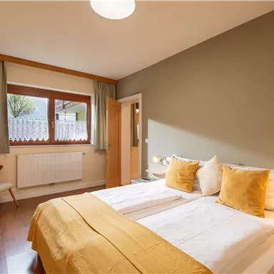A cozy bedroom with a double bed and colorful pillows. The room is bright and has large windows with a view outside.