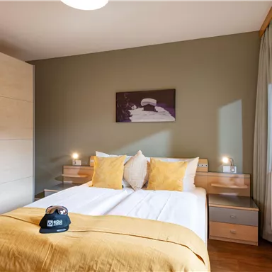 A modern bedroom with a double bed and yellow pillows. The room has light furniture and a large window with a view outside.