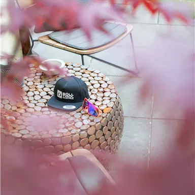 A black hat lies on a round table made of wooden logs. In the background, a chair and green plants can be seen.