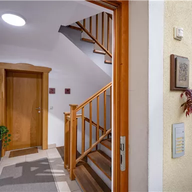 An inviting hallway with a wooden staircase and a warm light source. On the left, there is an entrance door and on the right, a decorative plant area.