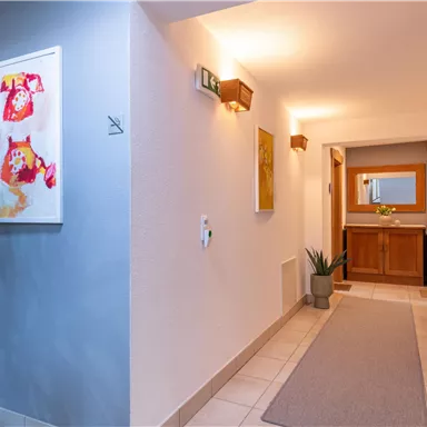 A bright hallway with modern artworks on the walls. On the right side, there is a wooden cabinet with plants and a mirror.