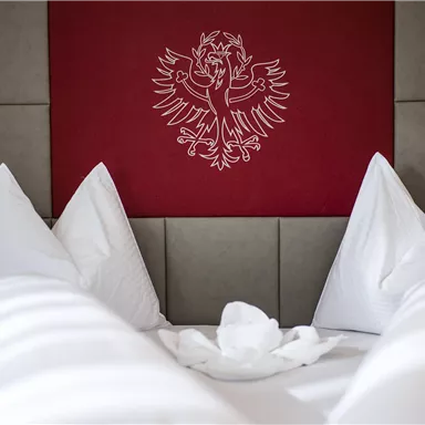 A cozy room with a large bed and many soft pillows. A stylized coat of arms is depicted on the wall.