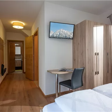 Ein helles Zimmer mit zwei Einzelbetten, einem Schreibtisch und einem Fernseher an der Wand. Die Holzverkleidung und die Vorhänge sorgen für eine gemütliche Atmosphäre.