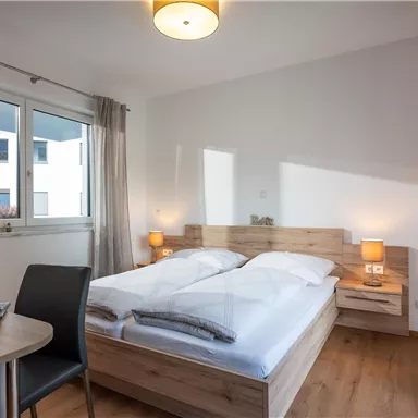 Ein modernes Schlafzimmer mit einem Doppelbett und Holzmöbeln. Der Raum ist hell und hat große Fenster für natürliches Licht.