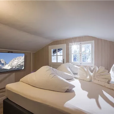 A cozy bedroom with a large bed and decorative pillows. A television hangs on the wall, while bright windows illuminate the room.