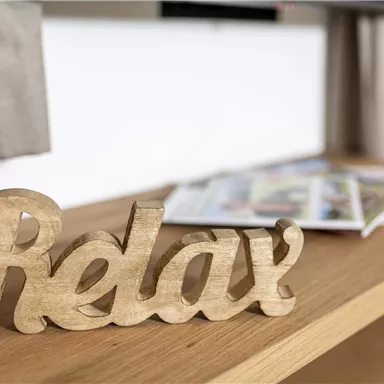 A decorative wooden sign with the inscription "Relax" is standing on a wooden table. In the background, there are some magazines.