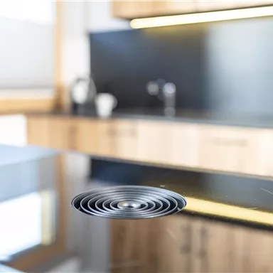 A modern kitchen counter made of black stone with a round hood. In the background, wooden cabinets and kitchen utensils are visible.