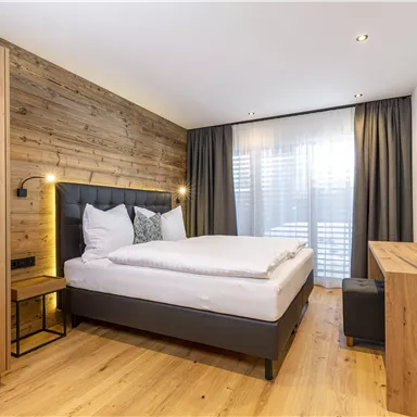 A modern bedroom with wooden paneling and a large window. It is stylishly furnished with a cozy bed and a wooden table.