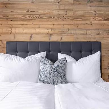A cozy bedroom with a tall leather headboard and numerous white pillows. The wooden wall gives the surroundings warmth and style.