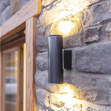 A modern outdoor lamp made of black metal on a stone wall. The light shines softly and creates a pleasant atmosphere.