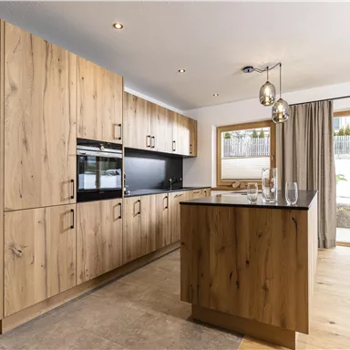 A modern kitchen with wooden cabinets and elegant lighting fixtures. Large windows provide plenty of daylight and a view outside.