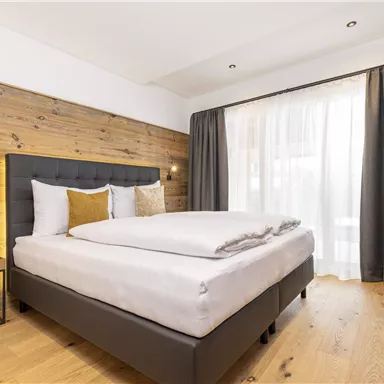 A modern bedroom with a comfortable bed and wood panels on the walls. Large windows let in plenty of daylight, and the curtains are elegantly draped.