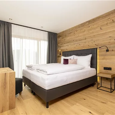 A modern bedroom with wood paneling and a large window. The room is bright and cozily furnished, featuring a comfortable bed and elegant furniture.