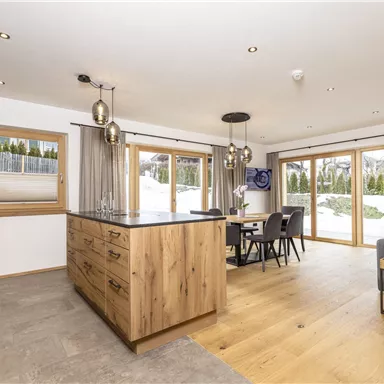 A modern kitchen with wooden cabinets and a large kitchen island. Large windows provide a view of the snowy landscape outside.