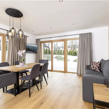 A modern living room with a dining table, chairs, and a cozy couch. Large windows offer a view of the snow-covered outdoor landscape.