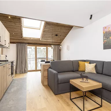 A modern living room with a comfortable gray sofa and a small table. The large windows let in a lot of light and there are artworks on the wall.
