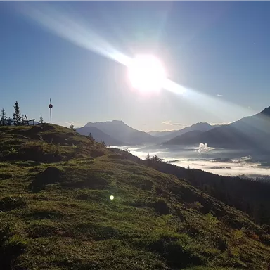 Eine malerische Berglandschaft mit der Sonne, die über den Gipfeln aufgeht. Der Himmel ist klar und der Nebel liegt sanft im Tal.
