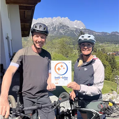 Ein Paar Radfahrer steht neben ihren Fahrrädern und hält ein Schild mit dem Logo "bett+bike". Im Hintergrund sind Berge und ein klarer blauer Himmel zu sehen.